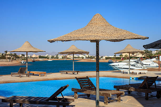 A beach with a pool and umbrellas. The umbrellas are all different sizes and are arranged around the pool. The pool is surrounded by chairs and a few boats are docked in the water